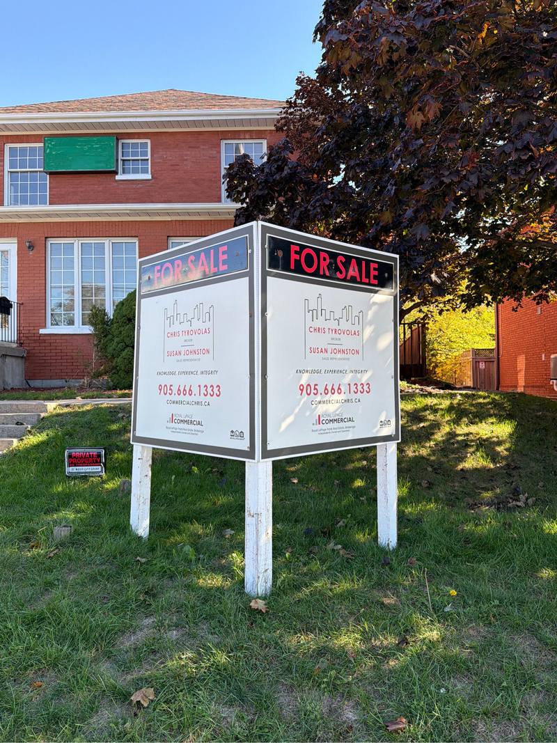 A large double sided commercial for sale sign on three wooden posts in front of a brick building, printed on both faces.